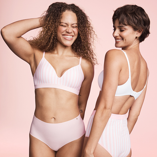 Two women standing side by side, one in pink underwear and the other in white underwear, smiling at the camera.